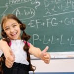 Smiling girl giving thumbs up in front of a chalkboard with math equations, wearing a white top.