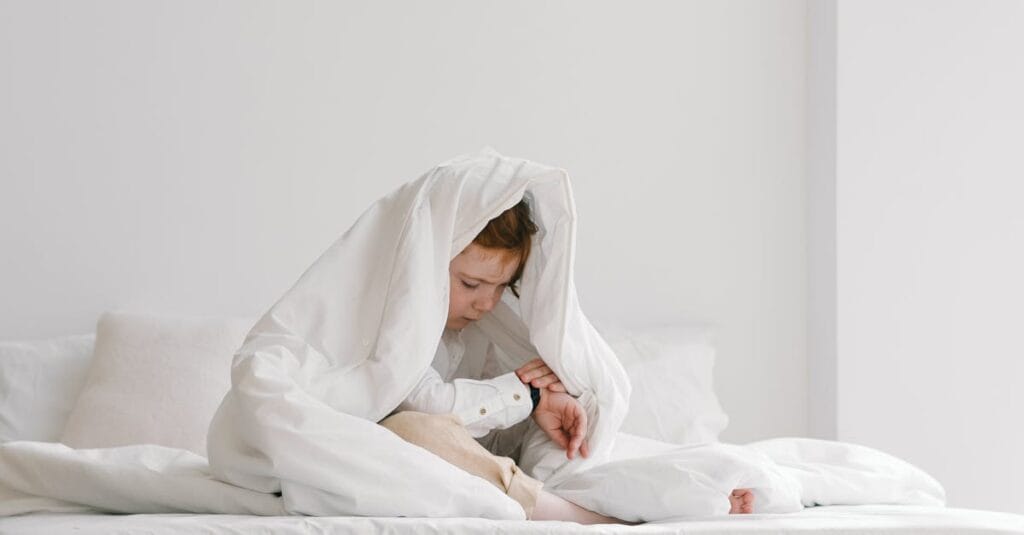 A young child sitting playfully under a white duvet on a bed in a bright room.