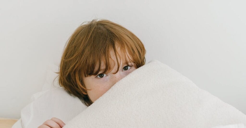 A young child with red hair peeks over a soft white blanket in a cozy indoor setting.