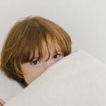 A young child with red hair peeks over a soft white blanket in a cozy indoor setting.