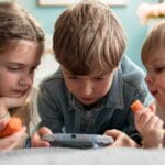 Three kids enjoying video games and snacking on carrots indoors, showcasing childhood togetherness.
