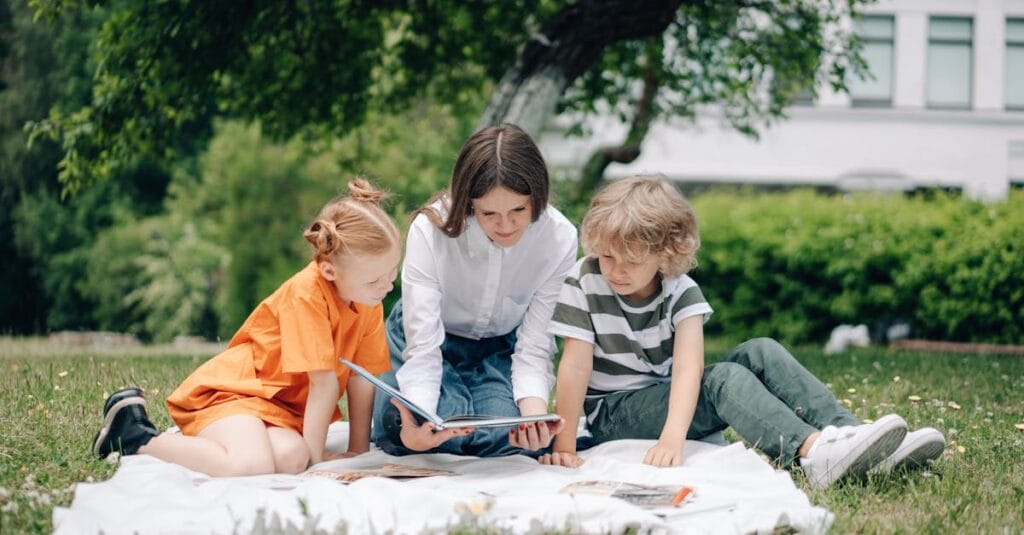 Three kids reading a book on a picnic blanket in a park under a tree.