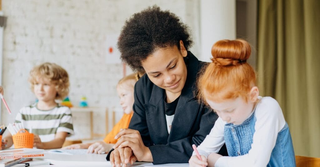 A teacher helps children with writing in a classroom setting, showcasing a diverse learning environment.