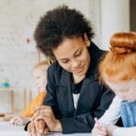 A teacher helps children with writing in a classroom setting, showcasing a diverse learning environment.
