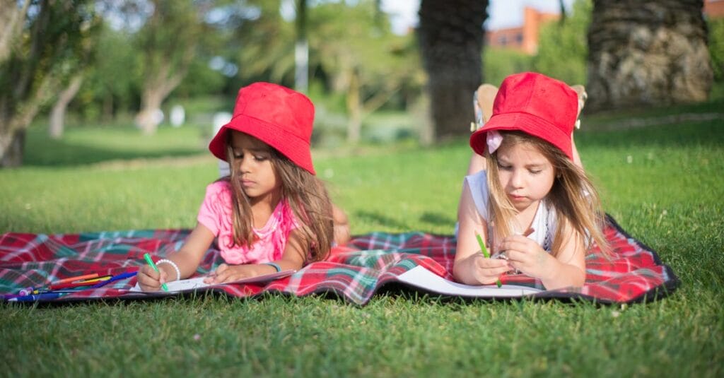 Children lying on a blanket, drawing in a park with red hats and sunny weather.