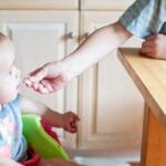 A cute baby in a high chair being fed by an older child in a bright kitchen setting.