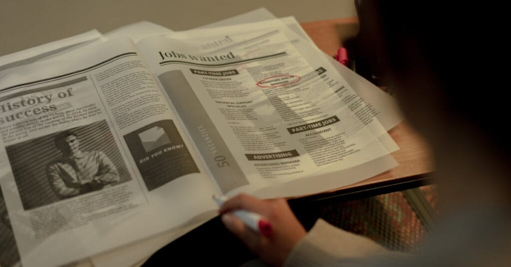 Person reviewing job listings in a newspaper, holding a pink marker for highlighting job ads.