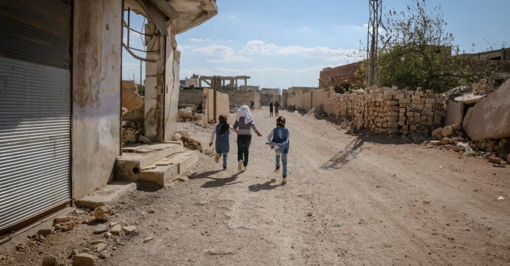 Kids run joyfully down a dirt road amidst war-damaged buildings in Idlib, Syria.