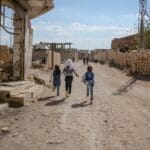 Kids run joyfully down a dirt road amidst war-damaged buildings in Idlib, Syria.