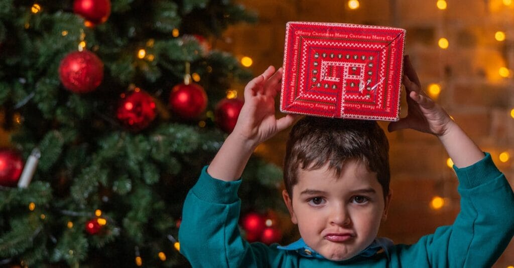 A young boy pouting while holding a Christmas gift above his head with a decorated tree in the background.