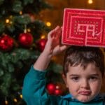 A young boy pouting while holding a Christmas gift above his head with a decorated tree in the background.