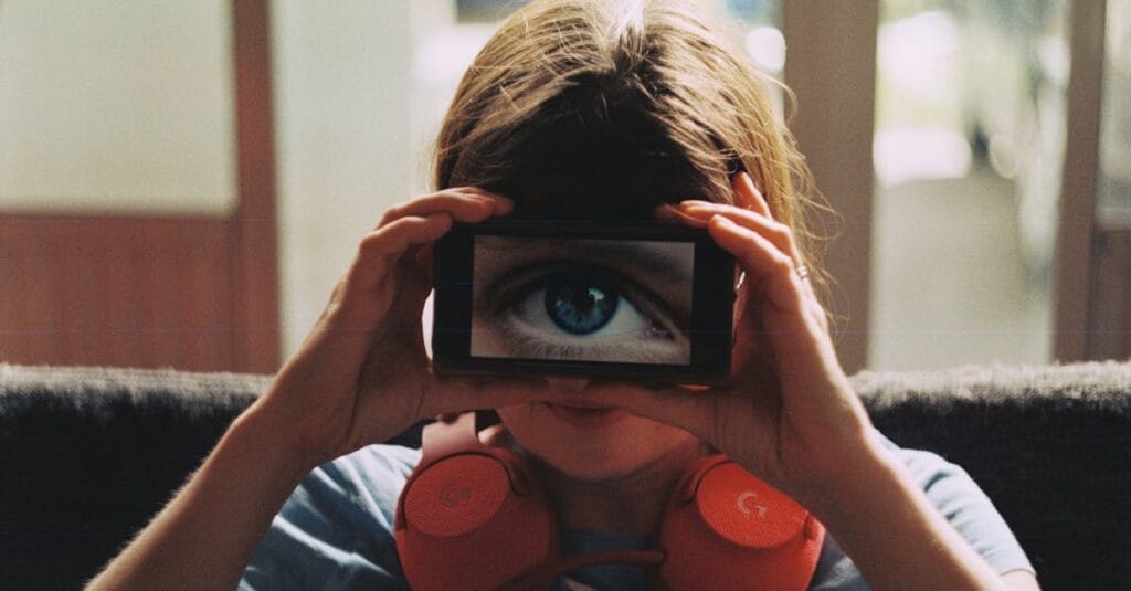 A person with headphones playfully holds a smartphone showing a large eye over their face.