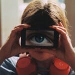 A person with headphones playfully holds a smartphone showing a large eye over their face.