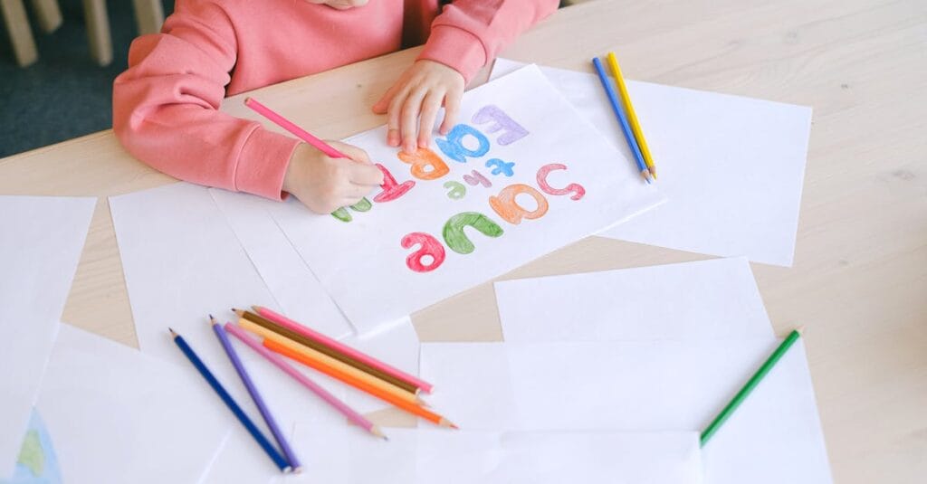 A child draws colorful letters on paper using colored pencils, showcasing creativity indoors.