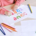 A child draws colorful letters on paper using colored pencils, showcasing creativity indoors.