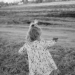 A young girl in a floral dress runs joyfully through a grassy field in black and white.
