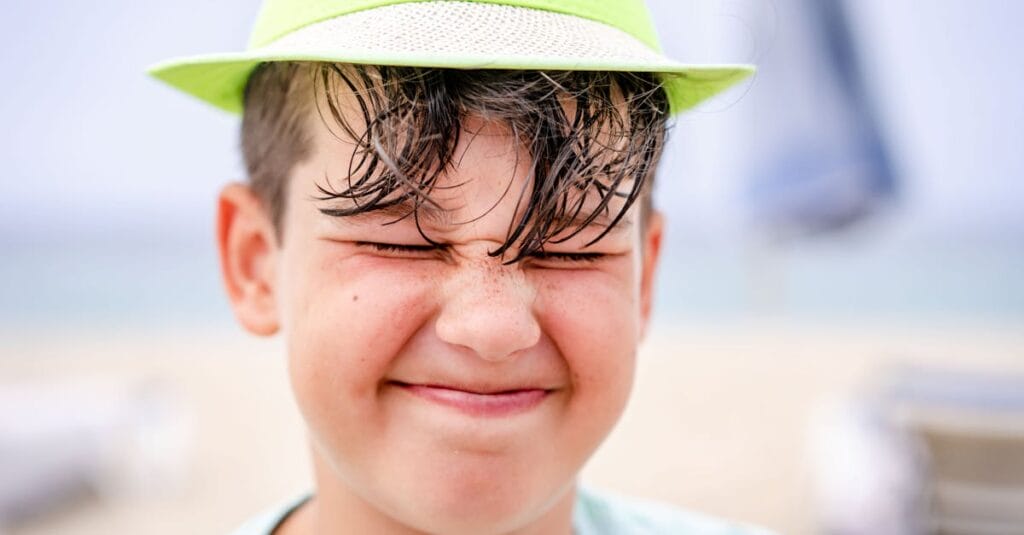 Close-up of a smiling boy with eyes closed and wearing a hat on a sunny beach day.