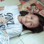 A smiling young girl lying on the floor surrounded by comic books, enjoying her reading time.