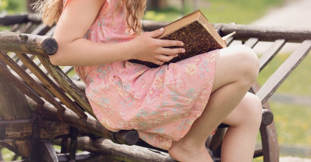 A young girl in a hat reads a book while sitting on a rustic wooden wagon outdoors.