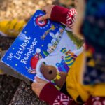 A child wearing colorful attire reads 'The Littlest Reindeer' book outdoors on a fall day.