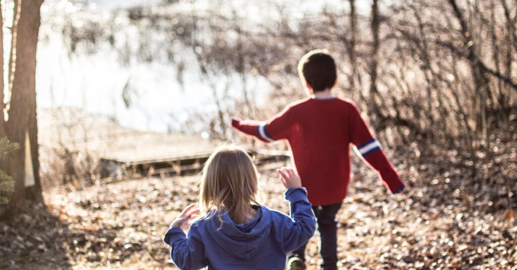 Two children running through a forest path by a lakeside on a sunny day.