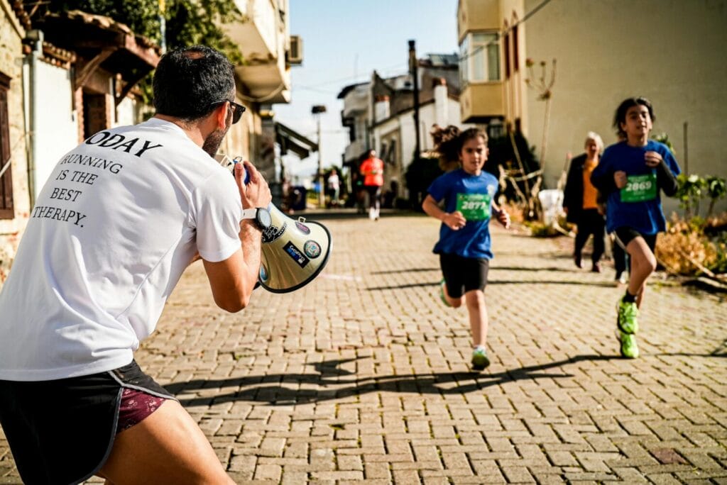Children running a street race while a man with a megaphone cheers them on.
