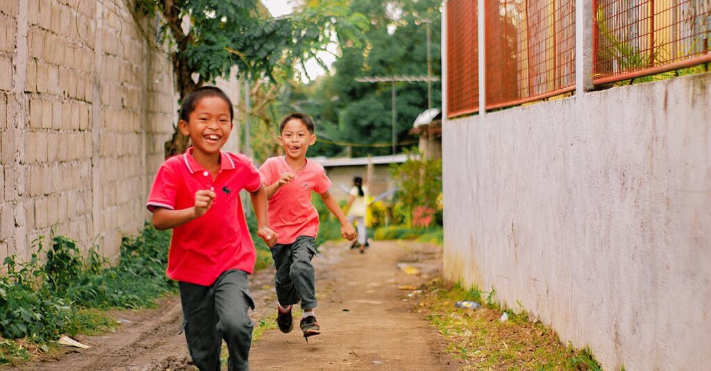 Two smiling children running joyfully on a sunny day through a rustic alleyway.