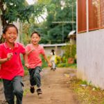 Two smiling children running joyfully on a sunny day through a rustic alleyway.