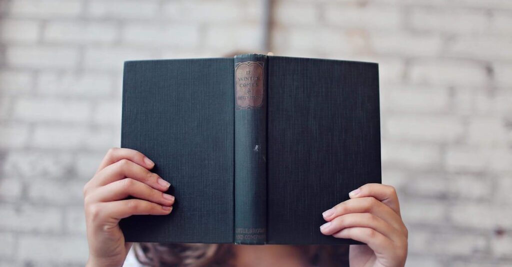 A woman holds an open book, reading in front of a brick wall.