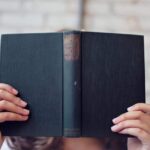 A woman holds an open book, reading in front of a brick wall.