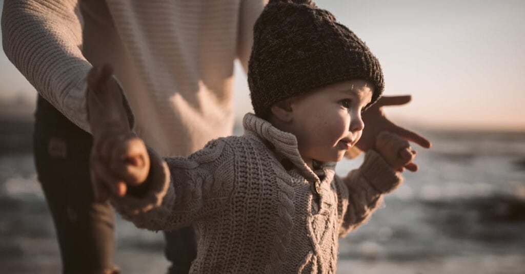 Adorable toddler in a knitted sweater and beanie enjoying a beach walk at sunset, holding hands with an adult.