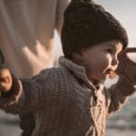 Adorable toddler in a knitted sweater and beanie enjoying a beach walk at sunset, holding hands with an adult.