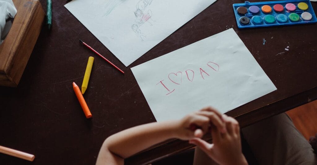 A child's handwritten note 'I ❤️ Dad' on paper surrounded by art supplies on a wooden table.