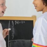 Two diverse girls discussing and solving a math equation on a chalkboard with enthusiasm.