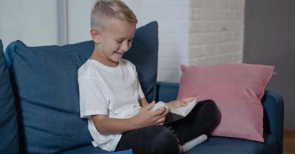 Cheerful blond boy reading a book on a cozy blue sofa indoors, smiling.