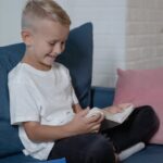 Cheerful blond boy reading a book on a cozy blue sofa indoors, smiling.