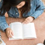 Overhead view of a woman reading a book outdoors on a sunny day, wearing a denim jacket.