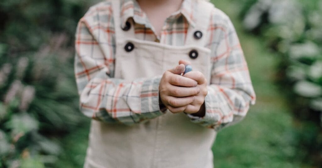 A young child holding a berry with greenery in the background, wearing a checkered shirt.