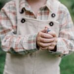 A young child holding a berry with greenery in the background, wearing a checkered shirt.
