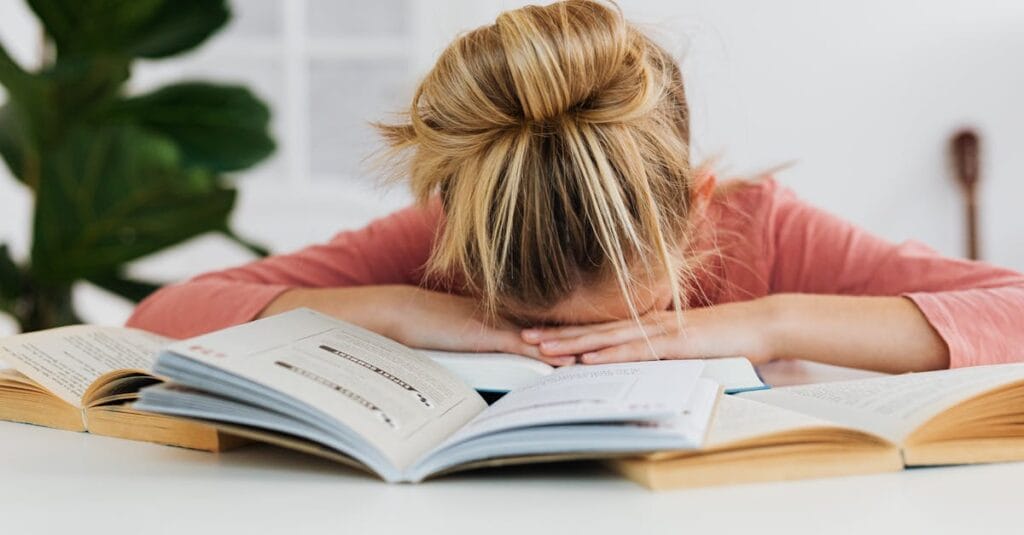 A young girl falls asleep on a desk surrounded by open books, illustrating study fatigue.