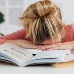 A young girl falls asleep on a desk surrounded by open books, illustrating study fatigue.