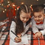 Two children writing letters on a cozy blanket by a decorated Christmas tree.