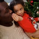 Father and daughter sharing a tender moment by the Christmas tree, enjoying the festive atmosphere.