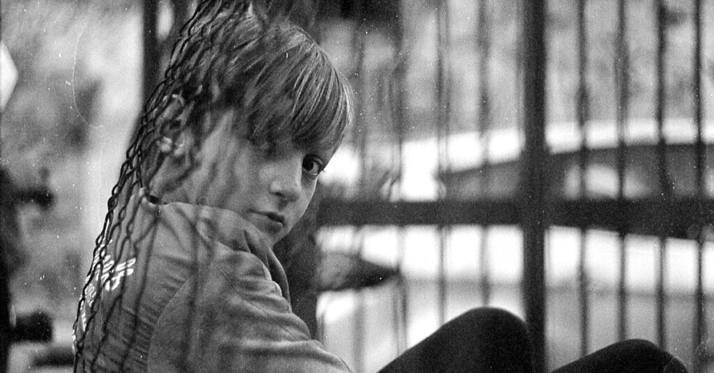 Black and white photo of a child sitting behind a wire cage, looking thoughtful.