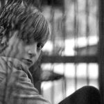 Black and white photo of a child sitting behind a wire cage, looking thoughtful.