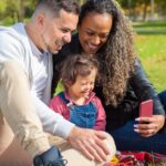 A happy family of three taking a selfie outdoors on a sunny day in the park.