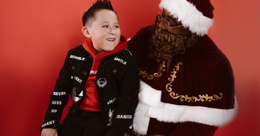 A child in a red hoodie smiles while sitting with Santa Claus in a studio setting.
