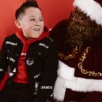 A child in a red hoodie smiles while sitting with Santa Claus in a studio setting.