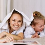 A joyful mother and daughter playfully read under a blanket on a cozy bed.
