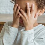 A young boy with curly hair covers his face with both hands, expressing distress or shyness in an indoor setting.